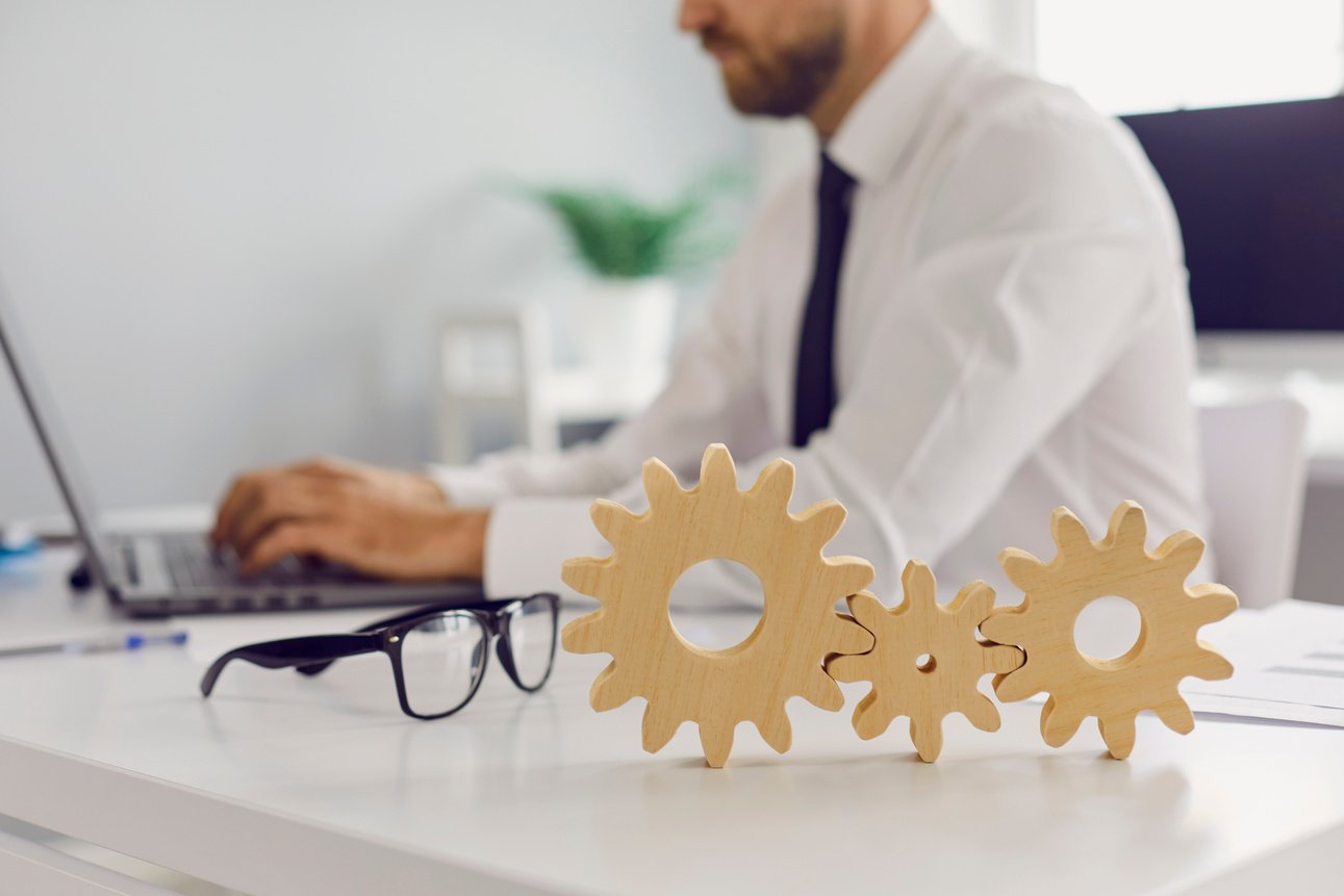 Three Cogwheels Joined Together Placed on Desk against Blurred Background of Busy Office Room